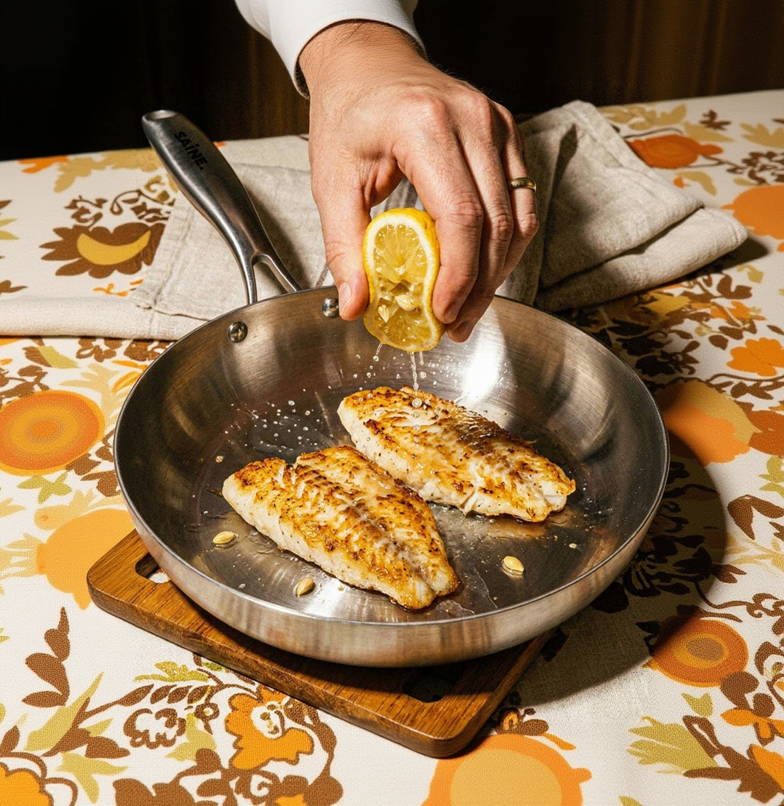 Filets de poisson dorés dans une poêle inox 26 cm SAINE avec une main qui presse un citron par-dessus, style vintage