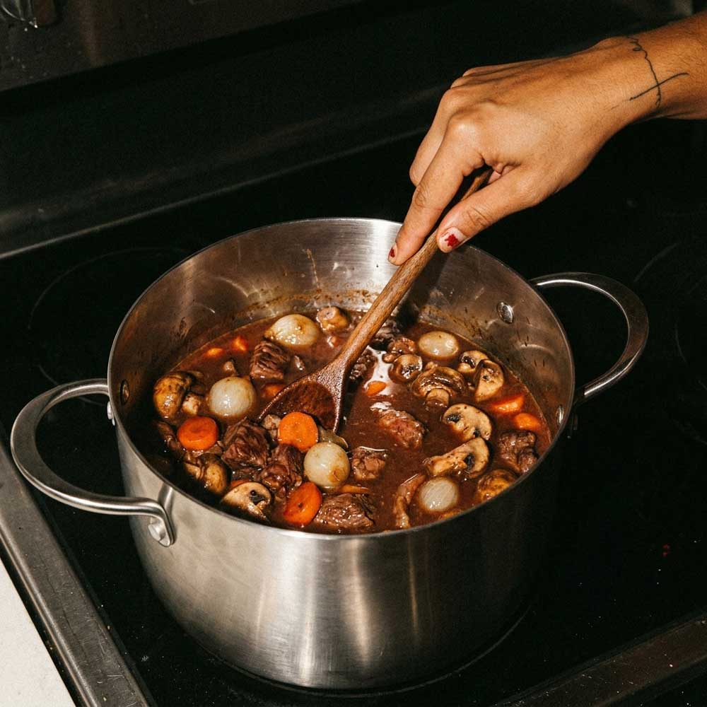 Boeuf bourguignon qui mijote dans une marmite inox SAINE, une main tatuée remuant la viande, champignons, carottes et oignons avec une spatule en bois