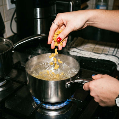 Main féminine versant des macaronis dans une casserole inox SAINE avec de l'eau bouillante sur feu de gaz vif