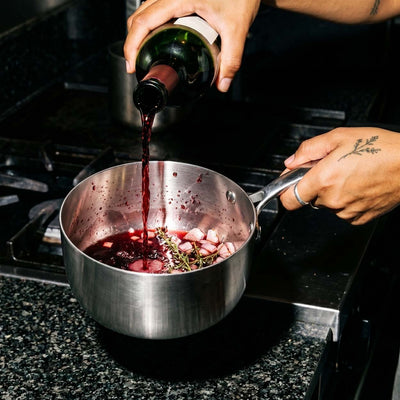 Mains versant du vin rouge dans une casserole inox SAINE pour réaliser une réduction avec des lardons et du thym sur une plaque de cuisson