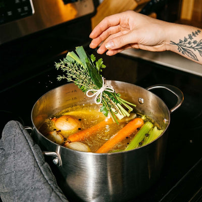 Main tatuée plongeant un bouquet garni dans une marmite inox SAINE avec un bouillon de légumes (carottes, oignon, poireau) sur plaque à induction