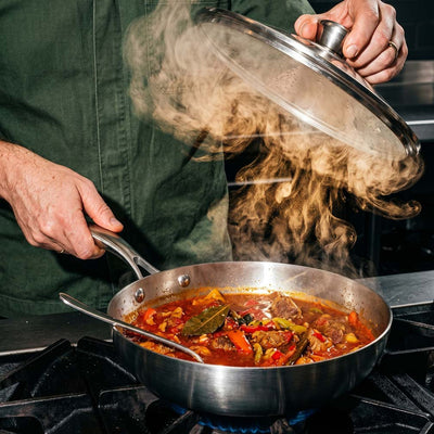 Homme soulevant le couvercle en verre d'une poêle inox SAINE sur laquelle mijote un ragoût de viande aux légumes sur feu de gaz, avec une vapeur spectaculaire