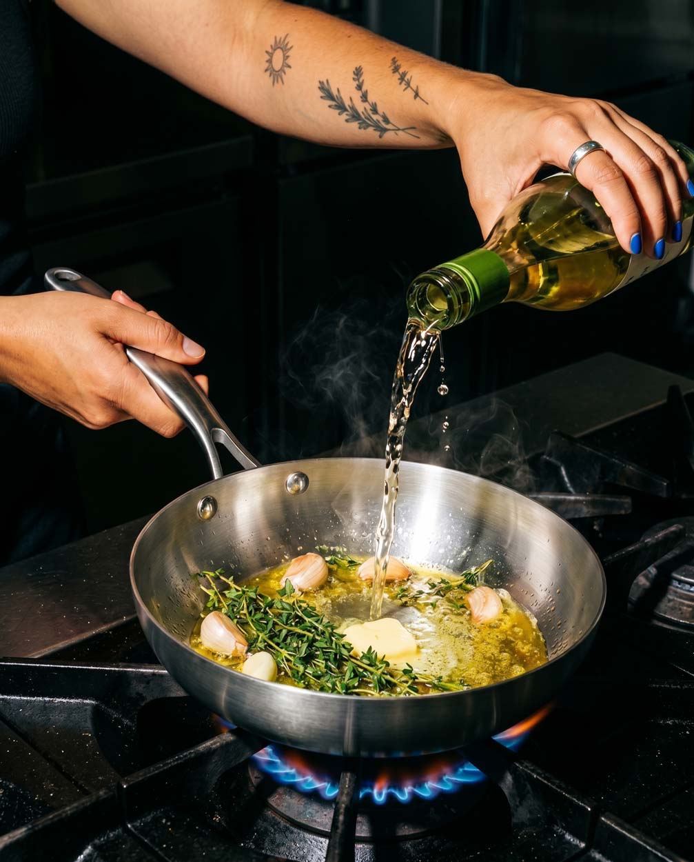 Main tatuée versant du vin blanc sur de l'ail, du thym et du beurre dans une poêle inox SAINE sur un feu de gaz à vive flamme