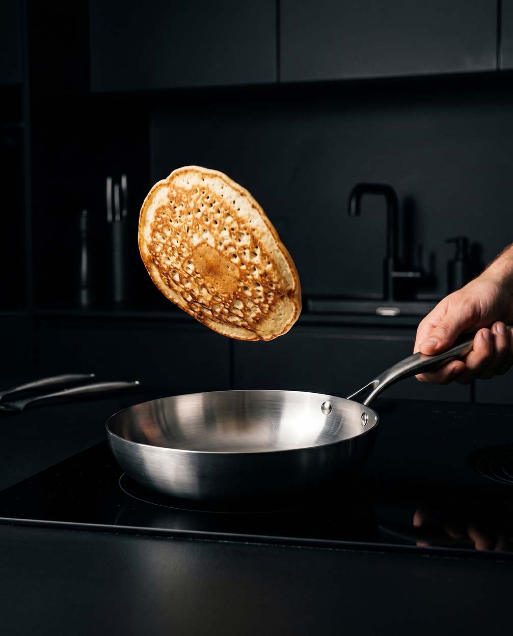 Crêpe dorée en l'air au-dessus d'une poêle inox 22 cm SAINE tenue à la main, sur une plaque à induction dans une cuisine noire