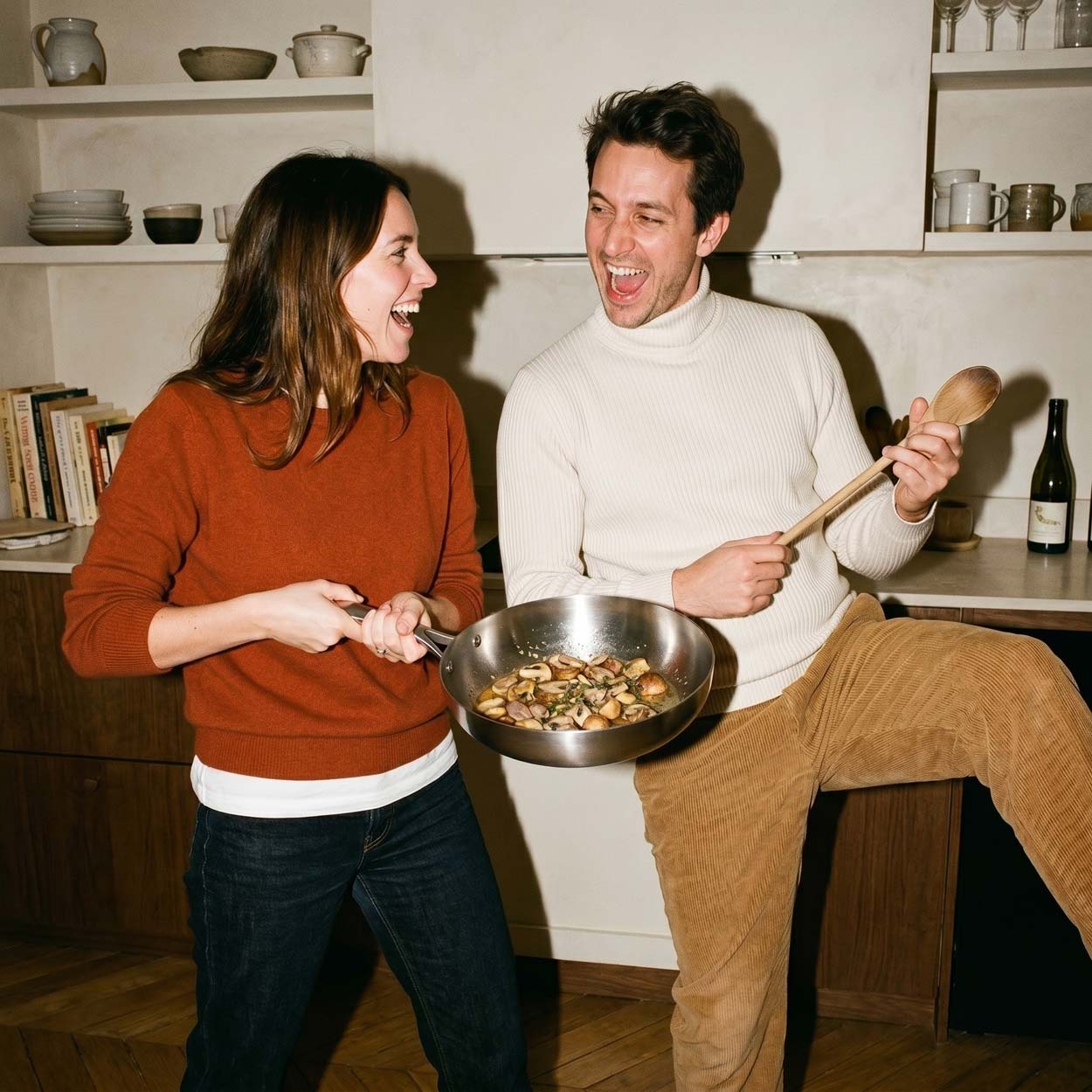 Couple comique en cuisine : la femme tient une poêle inox 24 cm SAINE avec des champignons sautés pendant que son compagnon mime une air guitar avec la spatule en bois
