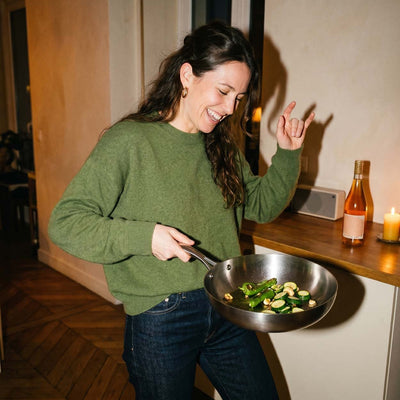 Femme joyeuse qui danse en faisant sauter des légumes (courgettes, haricots verts) dans une poêle inox 24 cm SAINE dans sa cuisine