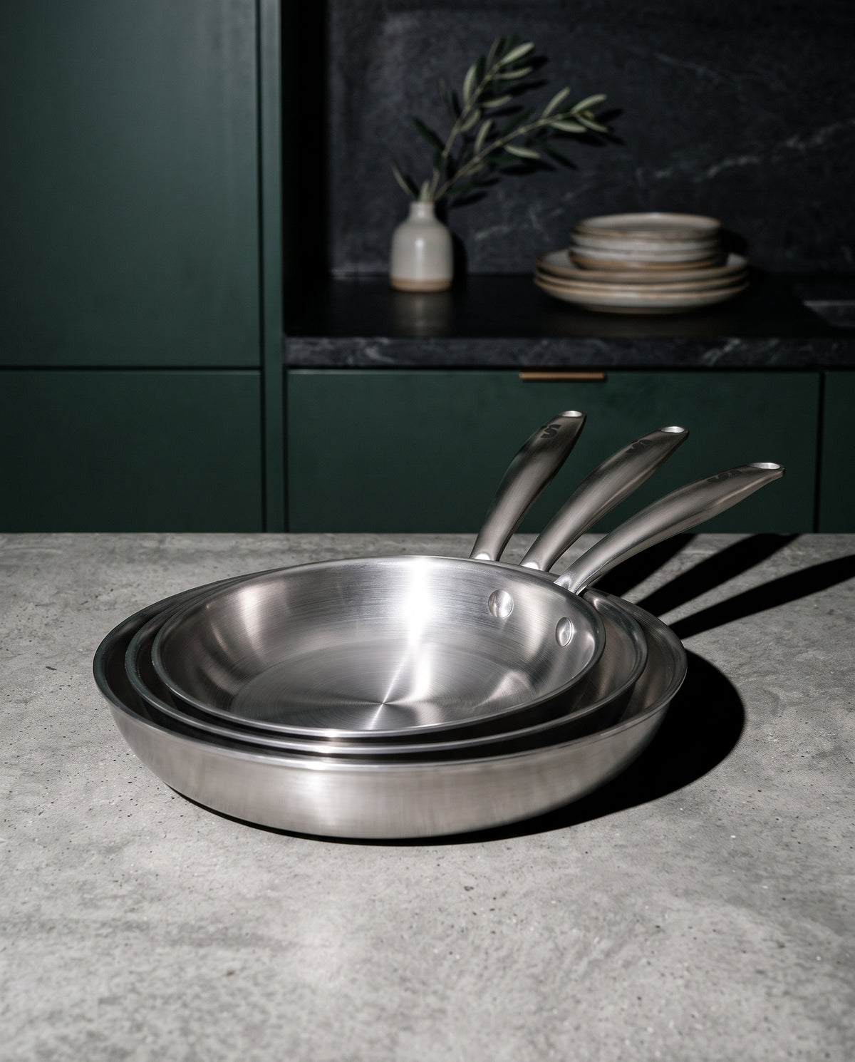 Trio de poêles inox SAINE sur un plan en béton, cuisine vert forêt