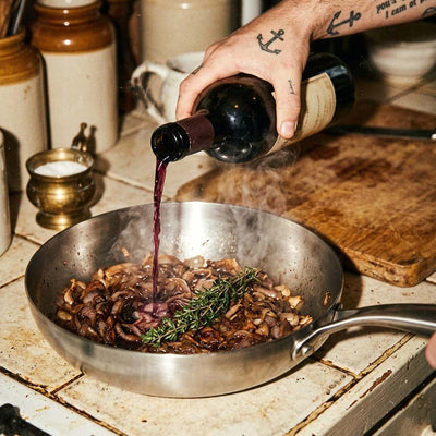 Main tatuée qui verse du vin rouge dans une poêle inox 22 cm SAINE avec des oignons caramlisés au thym, style vintage