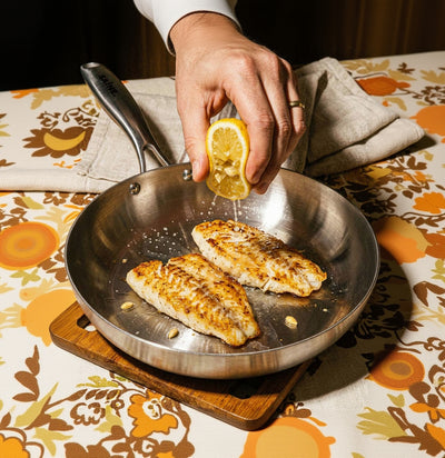 Filets de poisson dorés dans une poêle inox 26 cm SAINE avec une main qui presse un citron par-dessus, style vintage