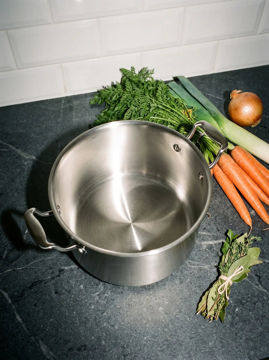 Marmite inox 24 cm SAINE sur un plan de travail en pierre, avec carottes, poireau, oignon et bouquet garni pour un pot-au-feu